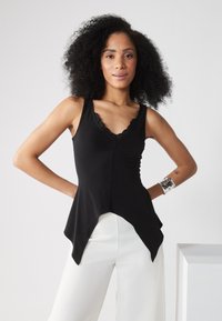 Top de tirantes negro ajustado con encaje en el escote; presenta un dobladillo asimétrico y acampanado. Combinado con pantalones blancos de pierna ancha.