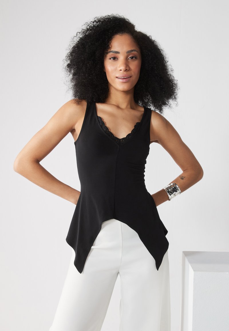 Top de tirantes negro ajustado con encaje en el escote; presenta un dobladillo asimétrico y acampanado. Combinado con pantalones blancos de pierna ancha.