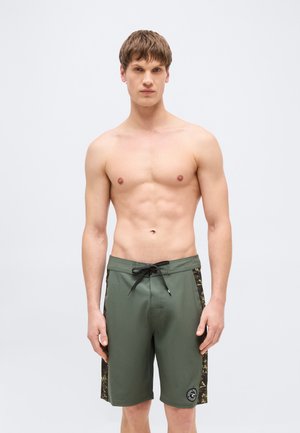 Giovane uomo con capelli castani corti che indossa pantaloncini da bagno verdi fino al ginocchio con coulisse nere e pannelli laterali mimetici, in piedi davanti a uno sfondo semplice.