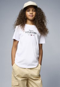 Bela bombažna majica s črnim napisom "Tommy HILFIGER" in letnico ustanovitve. V kombinaciji s bež cargo hlačami in bež kapo.