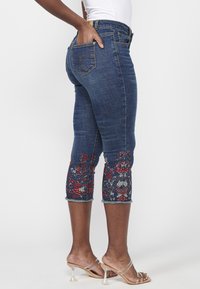 Dark denim cropped jeans with floral embroidery at the hem, featuring a fitted silhouette and frayed edges. Back pocket design visible.