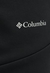 Λογότυπο της μάρκας Columbia σε λευκό πάνω σε μαύρο ύφασμα με ορατή υφή και διακριτική ραφή που διασχίζει διαγώνια το υλικό.