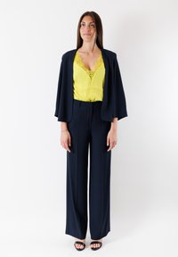 Blazer navy con maniche ampie sopra un canotta gialla brillante con bordo in pizzo, abbinato a pantaloni navy a vita alta e sandali neri.