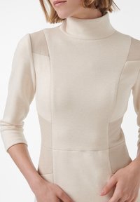 Beige coltrui jurk met textuur zijpanelen, met een getailleerd ontwerp en geribbelde accenten op de schouders en zijkanten.