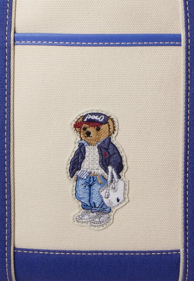 Kék-fehér színvilágú vászontáska, hímzett, hétköznapi ruhás medvével, aki egy táskát tart a kezében.