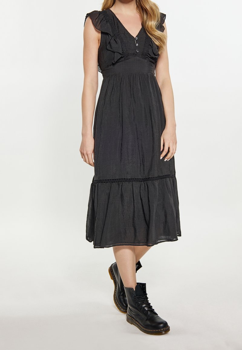 Robe midi noire avec des manches à volants, un décolleté en V et des boutons décoratifs. Elle présente un tissu texturé et une jupe à paliers froncée, accompagnée de bottes noires.
