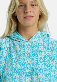 Poncho con capucha en color turquesa, con un patrón floral y de espirales, textura suave, que presenta un logotipo bordado en el pecho, diseñado para la comodidad y el uso diario.