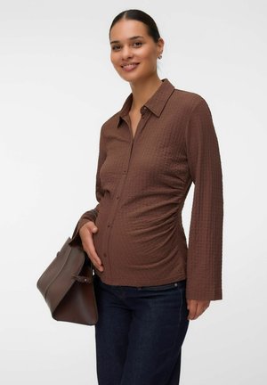 Donna incinta sorridente che indossa una camicia marrone a fantasia con bottoni e jeans scuri, che tiene una borsa marrone su uno sfondo bianco.