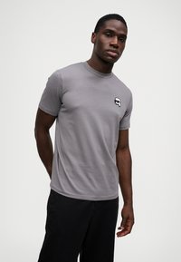 Γκρι κοντομάνικο μπλουζάκι με λαιμόκοψη, με μικρό κυκλικό λογότυπο στο αριστερό στήθος. Το ύφασμα φαίνεται λείο με ελαφριά ελαστικότητα.