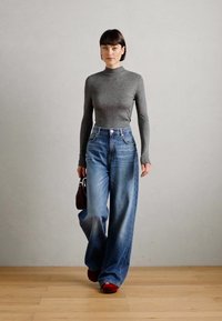 Top gris de manga larga, jeans azul de pierna ancha, zapatos rojos y un pequeño bolso oscuro. La modelo está de pie sobre un suelo de madera con un fondo gris.