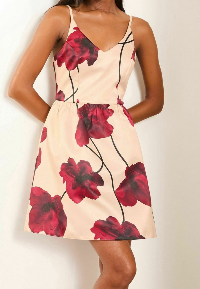 Korte bloemenjurk met een V-hals, dunne bandjes en een getailleerde taille. De stof is licht perzikkleurig met grote rode bloemen en zwarte stelen.