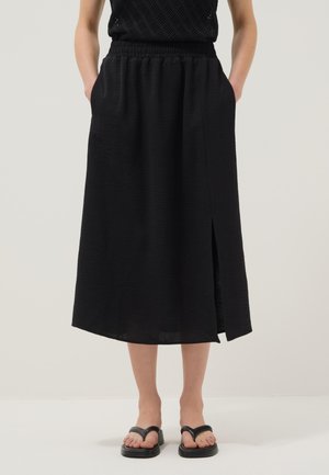 Donna con gonna midi nera con spacco laterale, top nero senza maniche e sandali neri bassi, in piedi davanti a uno sfondo bianco.