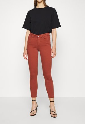 Woman wearing a black short-sleeve top, rust-colored skinny jeans with frayed hems, and black strappy high-heeled sandals standing against a white background.