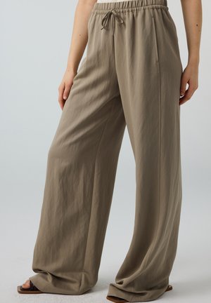 Loszittende beige broek met elastische tailleband en koordje, gedragen met bruine sandalen met vierkante neus op een neutrale achtergrond.