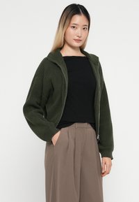 Giacca a maglia verde oliva con chiusura a zip, texture a coste e colletto alto, abbinata a un top nero e pantaloni beige.