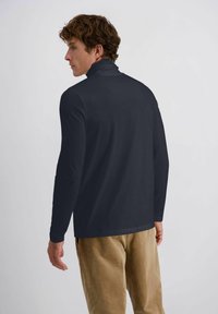 Mann mit lockigem braunem Haar, der einen dunkelblauen Langarmrollkragenpullover und beigefarbene Cordhosen trägt, seitlich vor einem schlichten Hintergrund stehend.