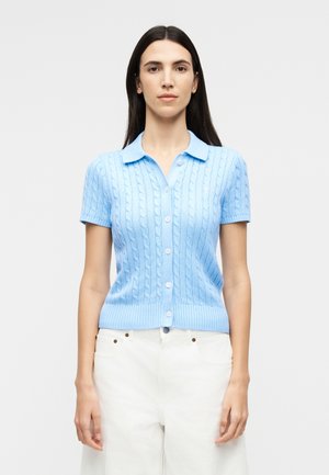 Kvinde med langt mørkt hår iført en lyseblå kortærmet kabelstrikket bluse med knapper og hvide bukser foran en ensfarvet baggrund.