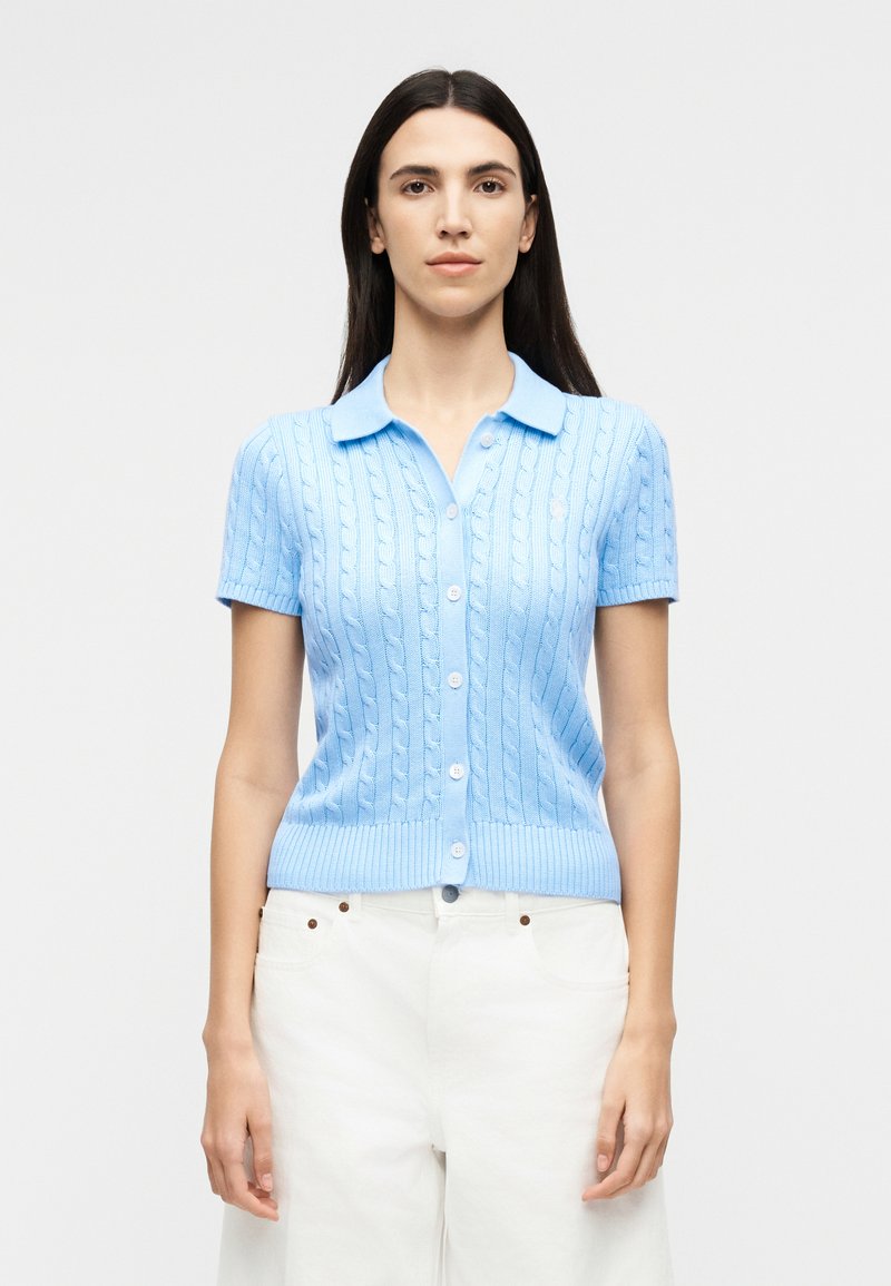 Sieviete ar gariem tumšiem matiem, valkājot gaiši zilu īss piedurkņu džemperi ar pogām un baltas bikses uz vienkārša fona.