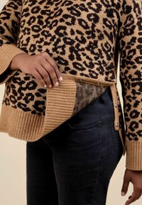 Un pull en imprimé léopard marron et noir avec des poignets et un ourlet côtelés, présentant une fente sur le côté et des accents de boutons, porté sur un jean foncé.