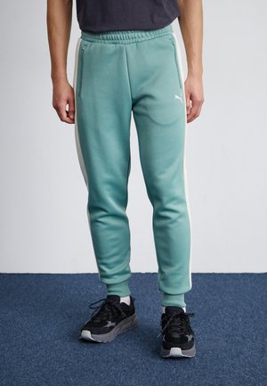 Hellblaue Jogginghose mit seitlichen weißen Streifen, elastischem Bund und vorderen Reißverschlusstaschen, kombiniert mit schwarzen Turnschuhen mit grauen Akzenten.