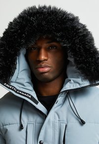 Šviesiai pilkas pūkinis švarkas su juodu kailiniu kapisonu, turintis priekinės kišenes, raištelius ir "Zavetti, Canada" etikete. Minkšta tekstūra.