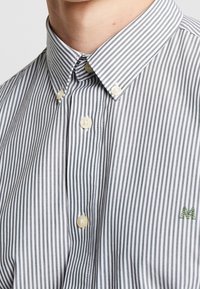 Camicia a righe con bottoni, caratterizzata da righe grigie e bianche alternate, un colletto con bottoni e un piccolo logo verde ricamato sul petto.