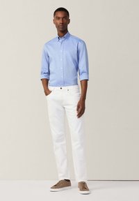 Camicia azzurra chiara con maniche arrotolate, jeans bianchi e sneakers marroni. La camicia presenta un colletto e un design a vestibilità classica.
