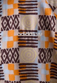 Adidas-mönstrad tyg har geometriska former i orange, blått och brunt på en beige bakgrund, med en vit logotyp broderad.