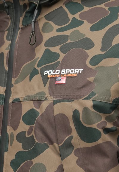 Kamuflāžas jaka audums ar izšūtu "Polo Sport Ralph Lauren" logotipu un nelielu Amerikas karodziņu pie rāvējslēdzēja un auklas cilpas.
