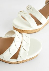 Sandales blanches à talon compensé avec trois brides texturées au design festonné, une semelle de type liège et un dessus en fausse cuir lisse.