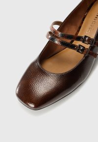 Braune Lederschuhe mit texturierter Oberfläche, quadratischer Zehenform und zwei glatten schwarzen Riemen mit Schnallen über dem Spann.
