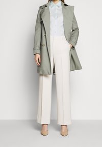 Trench coat verde chiaro sopra una camicia azzurra con bottoni, abbinato a pantaloni bianchi a vita alta e larghi e tacchi beige.