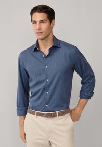 Herenoverhemd met knoopsluiting, donkerblauwe stof met een gestructureerde afwerking, lange mouwen opgerold bij de manchetten, witte knopen, gecombineerd met lichtbeige broek en een brown leren riem.