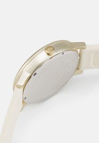Guldtonad armbandsur med en slät metallisk boett, glasbak och beiget silikonarmband med en texturerad yta och cirkulär design.
