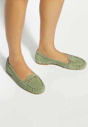 Jambes portant des mocassins en daim vert avec un détail mors d'or, debout sur une surface blanche.