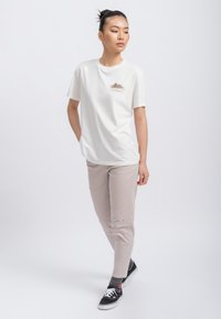 Witte katoenen T-shirt met een kleine grafische afbeelding op de voorkant, gecombineerd met lichtbeige broek en zwarte sneakers. Eenvoudige, casual pasvorm.