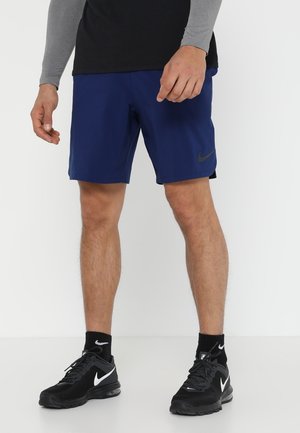 Mand iført blå Nike-shorts, sorte Nike-sneakers og sorte sokker, stående mod en hvid baggrund.