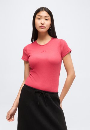 Mladá žena s dlhými čiernymi vlasmi nosí priliehavé červené tričko GAP a čiernu sukňu s vysokým pásom a sťahovacou šnúrkou na jednoduchom pozadí.