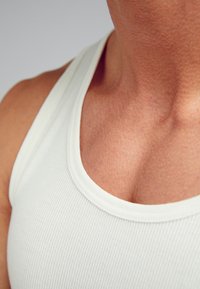 Vit ribbad linne med rund halsringning, som visar struktur och en slät yta. Fokusera på kragen och övre bröstområdet.