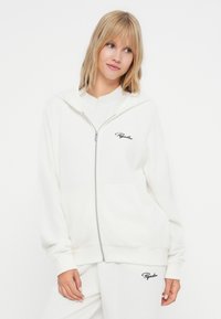 Bijela zip-up dukserica od rebraste tkanine s kapuljačom, s bočnim džepovima i crnim izvezenim logotipom na lijevoj strani prsnog koša.