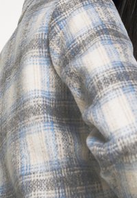 Gros plan d'une veste en tissu à carreaux doux gris, blanc et bleu clair portée par une personne aux longs cheveux foncés.