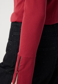 Červený dlhý rukáv s priliehajúcim strihom, ktorý má zips na manžetách. V kombinácii s čiernymi textúrovanými nohavicami s bodkovaným vzorom.