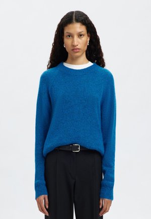 Heller blauer gestrickter Pullover mit Rundhalsausschnitt und langen Ärmeln, der eine weiche Textur aufweist. Kombiniert mit schwarzen Hosen und einem Gürtel.