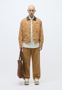 Brun canvasjakke med svart krage, hvit skjorte med knapper, brune vide bukser og en quiltet brun tote. Brukt med beige sneakers.