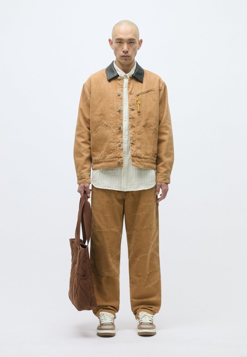 Braune Canvasjacke mit einem schwarzen Kragen, weißem Hemd, braunen weiten Hosen und einer gepolsterten braunen Tasche. Kombiniert mit beigen Turnschuhen.