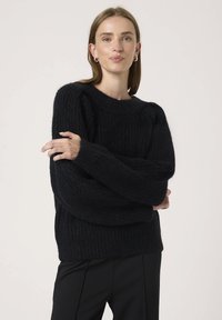 Pulover negru din tricot cu textură ribbinată, mâneci largi și decolteu rotund. Materialul pare moale și gros. Asortat cu pantaloni negri.