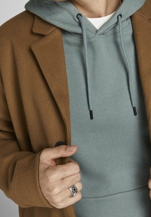 Bruine wolmix jas over een lichtblauwe hoodie. De hoodie heeft een kangoeroezak en koordjes met zwarte uiteinden. Ring om de hand.