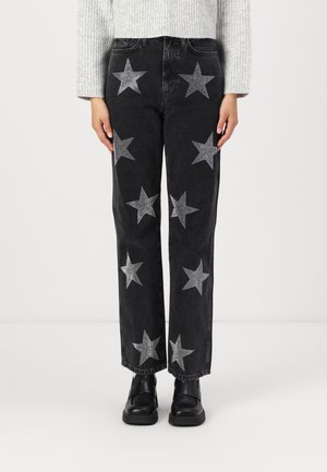 Jean noir avec des embellissements en étoile argentée, coupe droite, taille haute et poches, porté avec des chaussures noires et un pull gris clair.