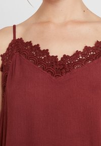 Top sin mangas color burdeos con un escote de encaje con forma de onda y una tela suave y texturizada. El diseño presenta un corte en forma de V en la parte delantera.