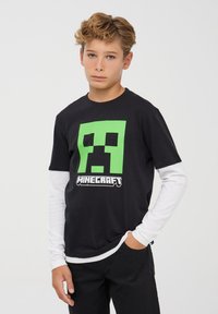 Czarna bawełniana koszulka z dużym zielonym nadrukiem twarzy creepera z Minecrafta oraz napisem "MINECRAFT". Nałożona na białą koszulę z długim rękawem.